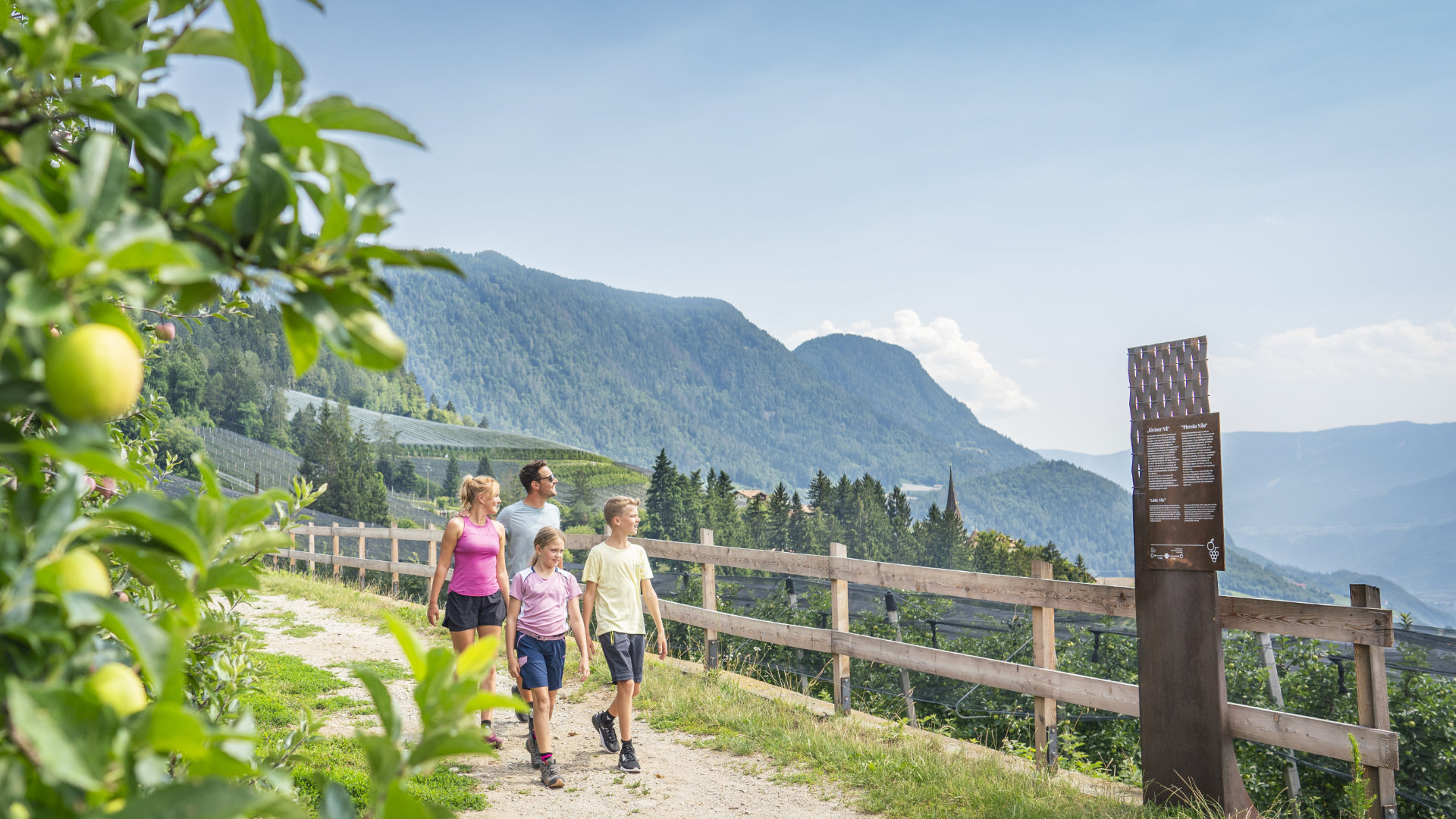 Familie beim Wandern zwischen Apfelgärten mit Panoramablick auf die Berge.