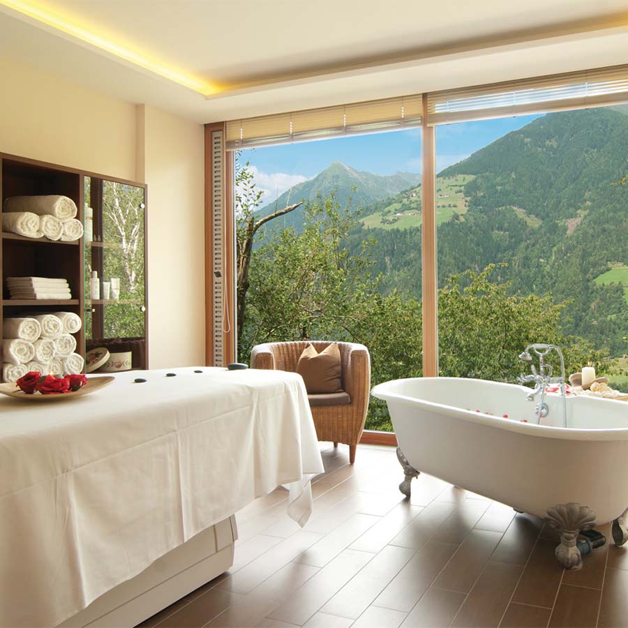 Spa-Kabine mit Massageliege und freistehender Badewanne vor einem großen Fenster mit Bergblick